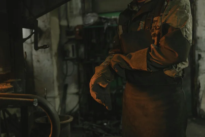 Osoba w skórzanym fartuchu i ciężkich rękawicach stoi przy rozżarzonym piecu w warsztacie.