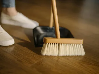 Miotła i szufelka na drewnianej podłodze, obok czyjeś białe buty.