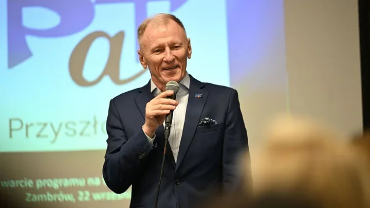 Mężczyzna w granatowym garniturze przemawia do mikrofonu podczas spotkania w sali konferencyjnej, w tle slajd z napisami.
