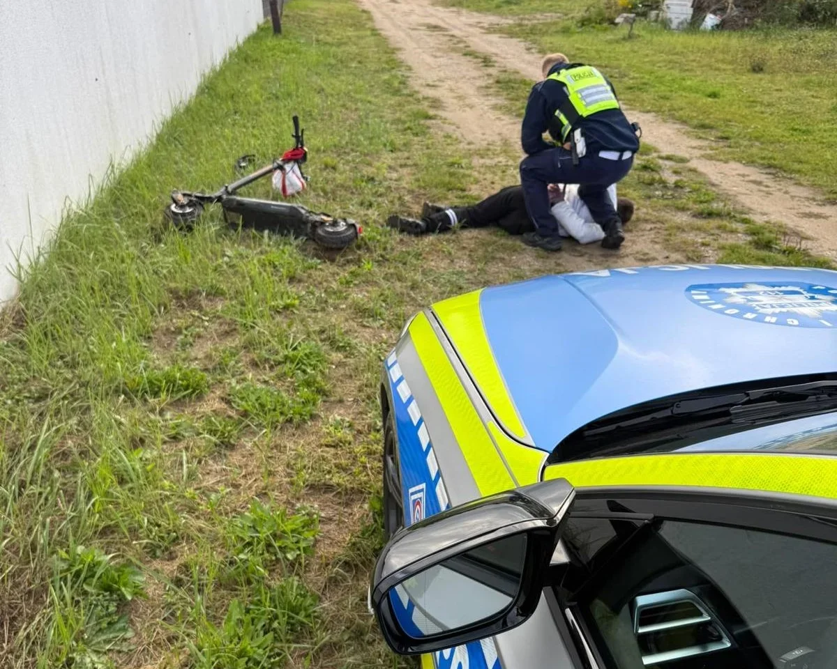 Policjant udziela pomocy leżącemu na trawie mężczyźnie obok przewróconej hulajnogi elektrycznej.