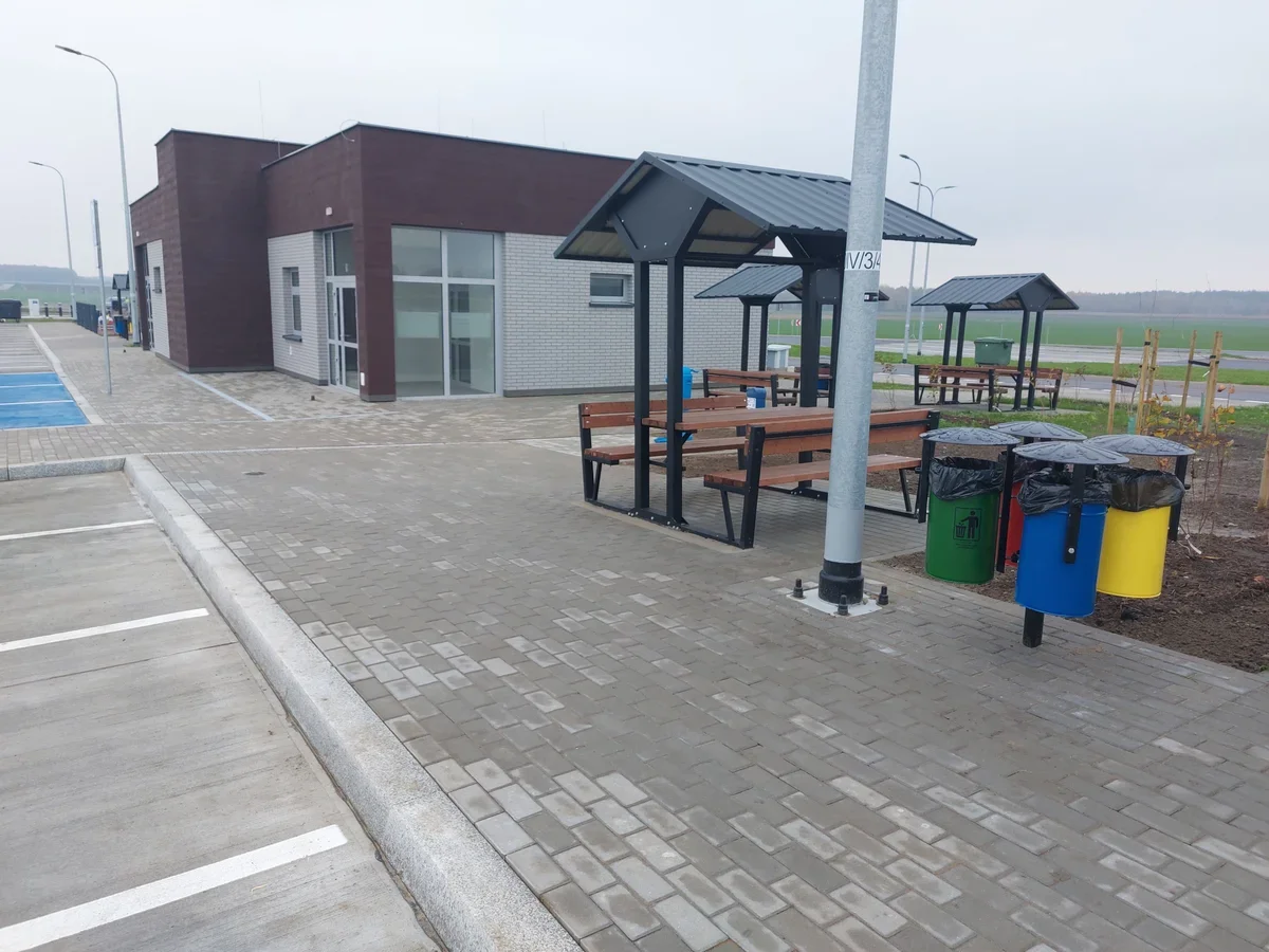 Nowoczesny budynek usługowy z wiatami przystankowymi, ławkami i koszami do segregacji odpadów na brukowanym placu przy parkingu.