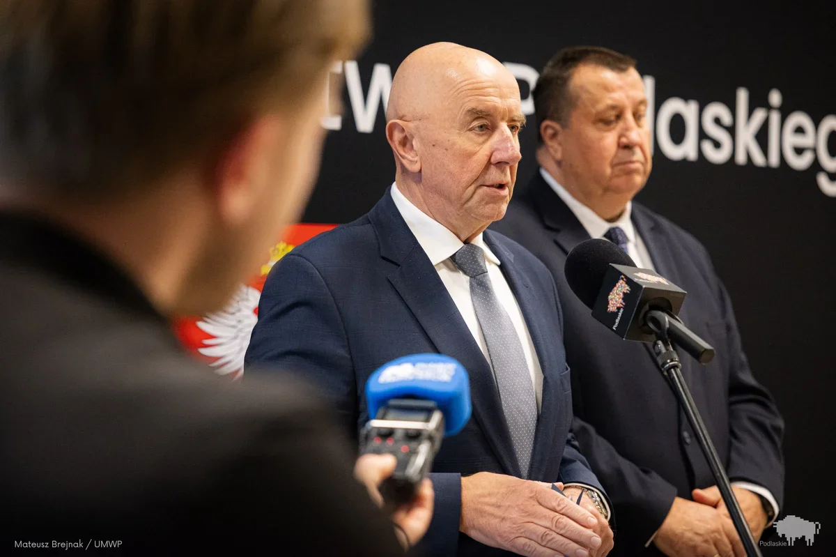 Dwaj mężczyźni w garniturach stoją podczas konferencji prasowej przed mikrofonami, jeden z nich przemawia.