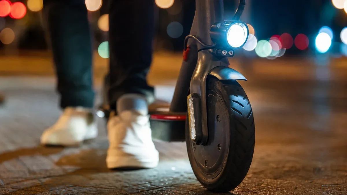 Przednie koło hulajnogi elektrycznej z włączonym reflektorem na brukowanej nawierzchni w nocy, obok stoją nogi osoby w białych butach.