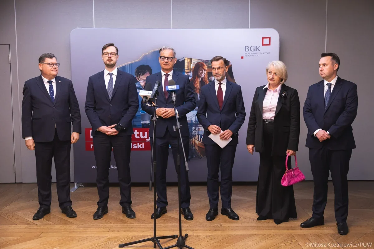 Sześć osób w formalnym stroju stoi podczas konferencji prasowej przed ścianką z logo BGK.