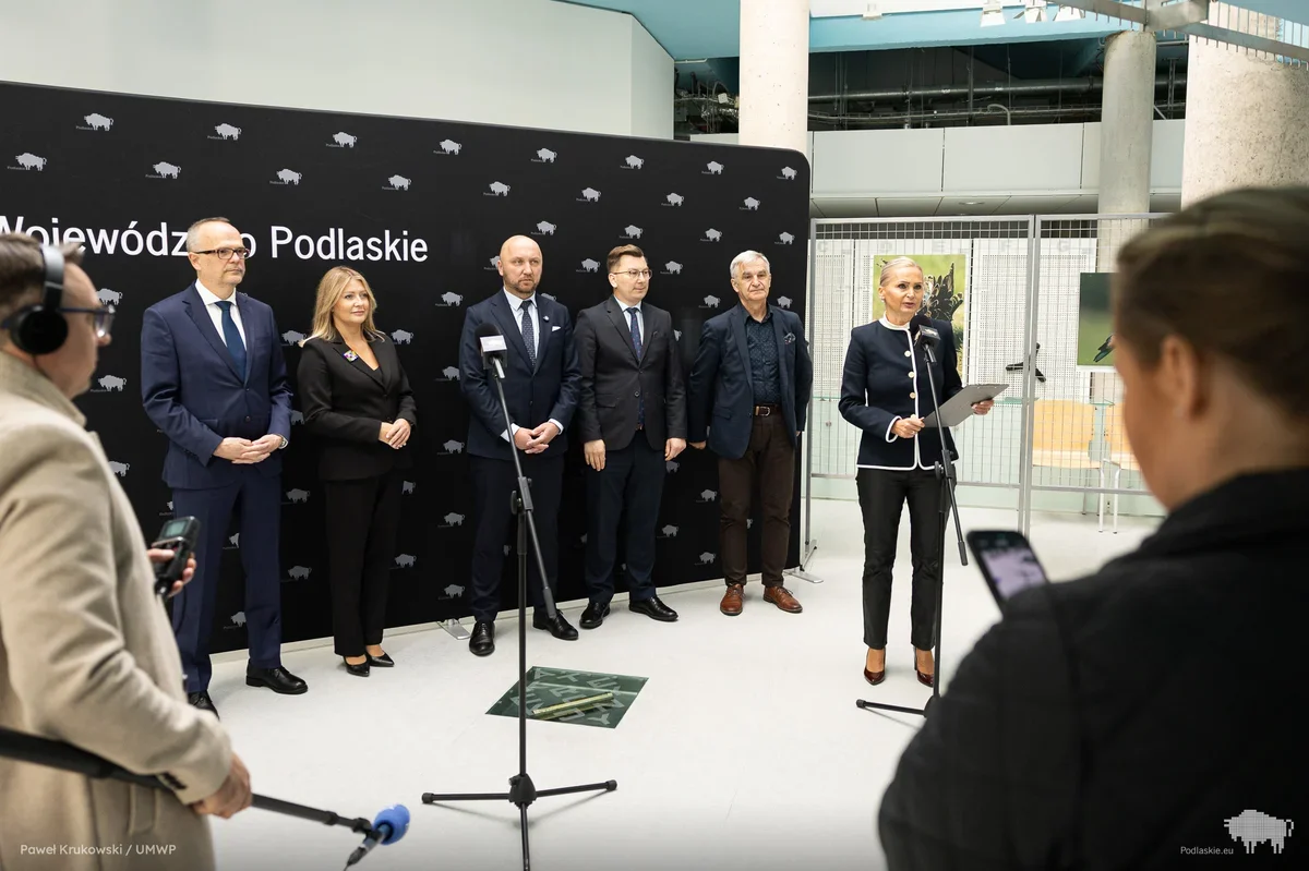 Grupa osób w eleganckich strojach stoi przed ścianką z napisem „Województwo Podlaskie” podczas konferencji prasowej w nowoczesnym wnętrzu.
