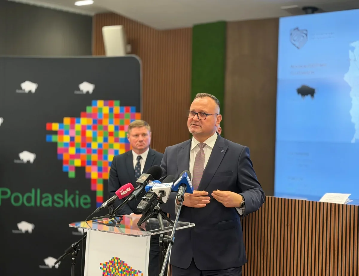 Mężczyzna w garniturze przemawia przy pulpicie z mikrofonami podczas konferencji prasowej w sali z logo województwa podlaskiego.