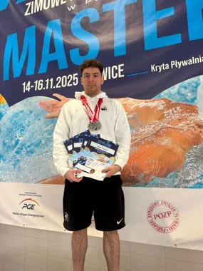 Mężczyzna z medalami i dyplomami pozuje na tle baneru zawodów pływackich Masters.