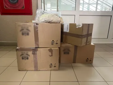 Cztery kartony opakowane taśmą stoją na posadzce w holu przy drzwiach, na jednym z nich leży worek z materiałem tekstylnym.