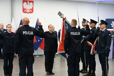 Funkcjonariusze Policji składają przysięgę podczas uroczystej ceremonii w pomieszczeniu z flagami Polski i Unii Europejskiej oraz herbem na ścianie.
