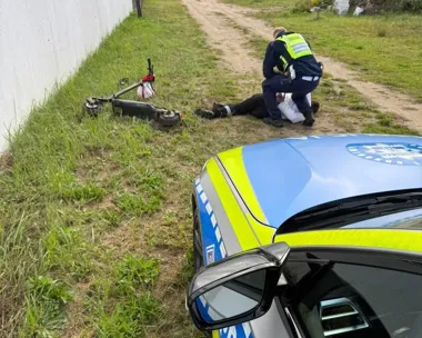 Policjant udziela pomocy leżącemu na trawie mężczyźnie obok przewróconej hulajnogi elektrycznej.