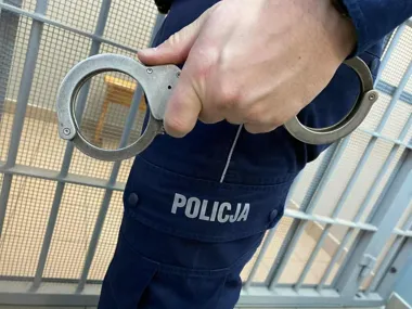 Funkcjonariusz policji w granatowym mundurze trzyma w dłoni metalowe kajdanki, w tle fragment ogrodzenia z siatki.