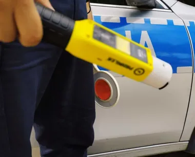 Funkcjonariusz w mundurze trzyma alkomat obok policyjnego radiowozu.