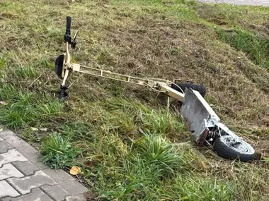 Rozbita, beżowa hulajnoga elektryczna leży na zarośniętej trawie przy brukowanym chodniku.