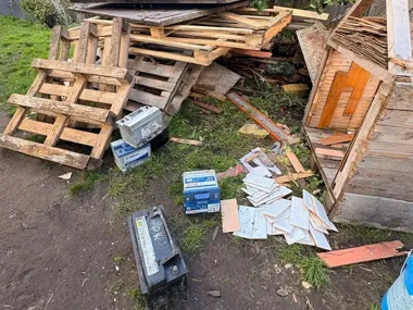 Zużyte akumulatory samochodowe, palety i drewniane odpady leżą na trawie i ziemi w nieuporządkowanym miejscu na zewnątrz.