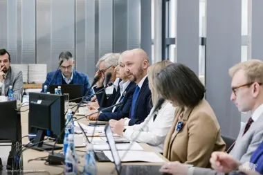 Uczestnicy oficjalnego spotkania siedzą przy stole konferencyjnym z monitorami i mikrofonami w nowoczesnym pomieszczeniu biurowym.