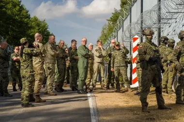 Grupa wojskowych i żołnierzy stoi przy ogrodzeniu z drutem kolczastym podczas wizyty lub inspekcji na granicy.