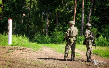 Dwóch żołnierzy w pełnym umundurowaniu stoi na leśnej drodze przy słupku granicznym.