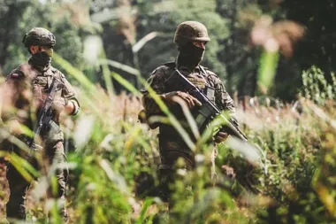 Dwaj żołnierze w kamuflażu i z bronią długą patrolują zarośnięty teren.