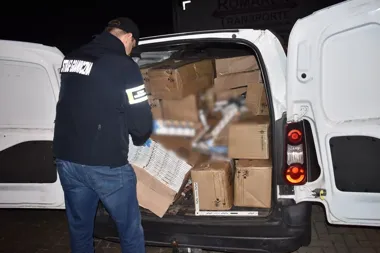 Funkcjonariusz Straży Granicznej przy białym furgonie wypełnionym kartonami i paczkami z papierosami podczas nocnej kontroli.