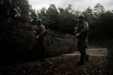 Dwóch żołnierzy w pełnym umundurowaniu i z bronią patroluje teren przy zasiekach z drutu kolczastego w lesie.