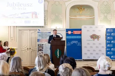 Mężczyzna przemawia z mównicy podczas uroczystości jubileuszu 70-lecia w reprezentacyjnej sali, w tle banery instytucji regionalnych i medialnych.