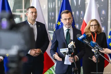 Mężczyzna w garniturze przemawia do mediów podczas konferencji prasowej, za nim stoją dwie osoby oraz flagi Unii Europejskiej i Polski.