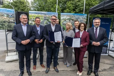 Grupa osób w strojach biznesowych pozuje na zewnątrz z otwartymi dokumentami w rękach, w tle plansze z wizualizacjami infrastruktury miejskiej i namiot z napise