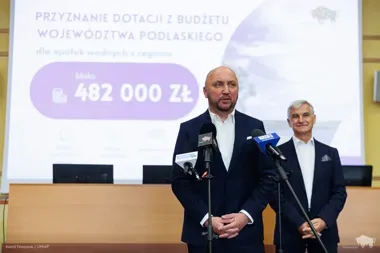 Dwaj mężczyźni w garniturach stoją przy mikrofonach podczas konferencji prasowej o przyznaniu dotacji dla spółek wodnych z województwa podlaskiego.
