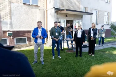 Grupa osób z identyfikatorami stoi na trawniku przed budynkiem mieszkalnym podczas zewnętrznego spotkania.