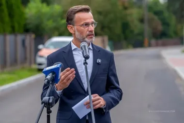 Mężczyzna w garniturze przemawia do mikrofonów na zewnątrz, na tle ulicy i zieleni.