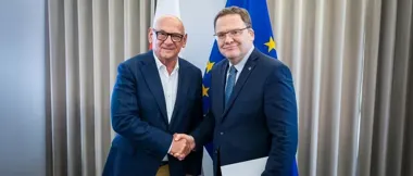 Dwaj mężczyźni w garniturach witają się uściskiem dłoni przed flagami, w tym flagą Unii Europejskiej.