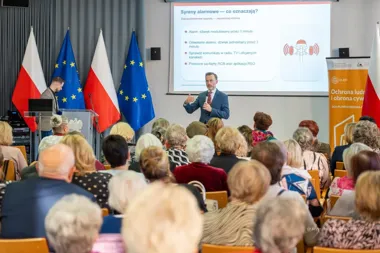 Mężczyzna w garniturze prowadzi wykład dla grupy seniorów w sali konferencyjnej o syrenach alarmowych, na tle flag polskich i unijnych.