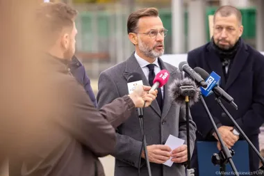 Mężczyzna w szarym płaszczu przemawia podczas konferencji prasowej na zewnątrz, otoczony mikrofonami mediów.