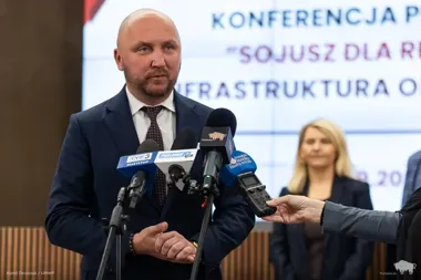 Mężczyzna w garniturze przemawia podczas konferencji prasowej do mikrofonów mediów regionalnych.