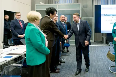 Mężczyzna w garniturze wita się uściskiem dłoni z kobietą w brązowym żakiecie podczas spotkania w sali konferencyjnej.