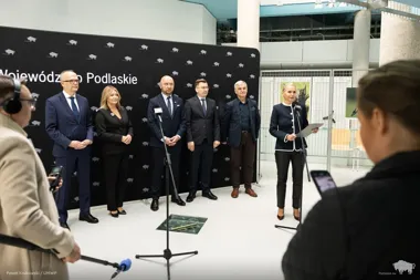 Grupa osób w eleganckich strojach stoi przed ścianką z napisem „Województwo Podlaskie” podczas konferencji prasowej w nowoczesnym wnętrzu.