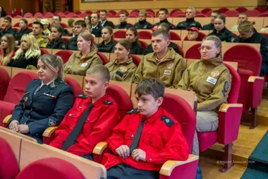 Uczestnicy w mundurach siedzą na czerwonych fotelach w auli podczas uroczystości lub konferencji.