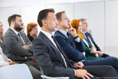 Uczestnicy w formalnym stroju siedzą w rzędach krzeseł podczas konferencji lub spotkania biznesowego w jasnej sali.