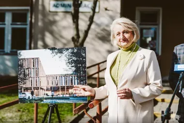 Kobieta w jasnym płaszczu stoi na zewnątrz obok wizualizacji nowego budynku na sztalugach, podczas wydarzenia prasowego przed budynkiem.