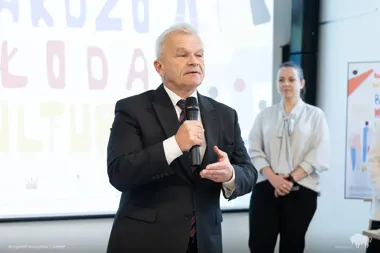 Mężczyzna w garniturze przemawia do mikrofonu podczas wydarzenia, w tle kobieta i slajd z napisami.