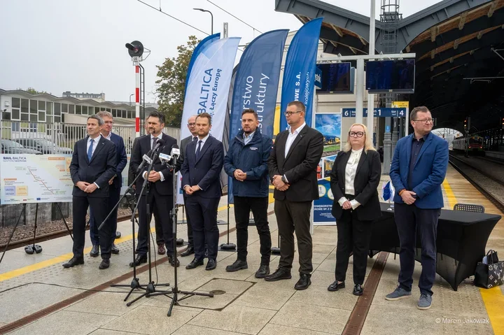 Grupa osób stoi na peronie kolejowym podczas konferencji prasowej dotyczącej projektu Rail Baltica.